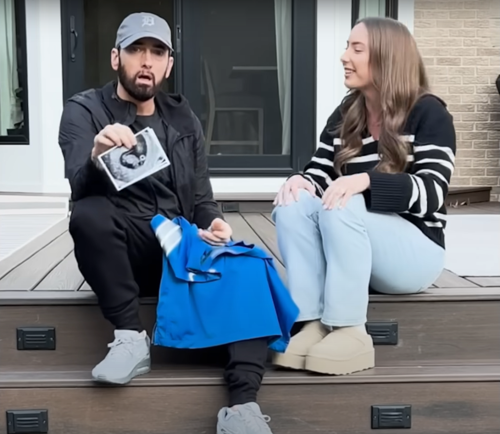 In the emotional music video for "Temporary," Eminem includes a touching scene where Hailie surprises her father with a blue jersey that reads “#1 Grandpa” alongside a sonogram of her unborn child. (Image Source: YouTube @eminem)