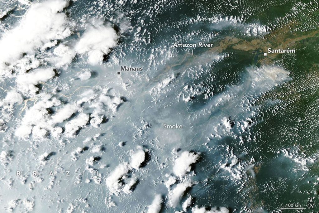 Thick gray smoke partially obscures the Amazon River and surrounding rainforest near the cities of Manaus and Santarém. Puffy white clouds rise above the smoke on the left side of the image.