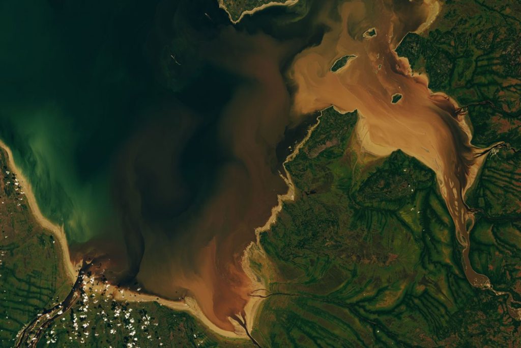 Dark brown water from the Nottaway, Harricanaw, and Moose rivers swirls into dark blue bay waters and mingles with plumes of lighter brown suspended sediment. Charlton Island and the much smaller Stag Rock are visible close to the shoreline.