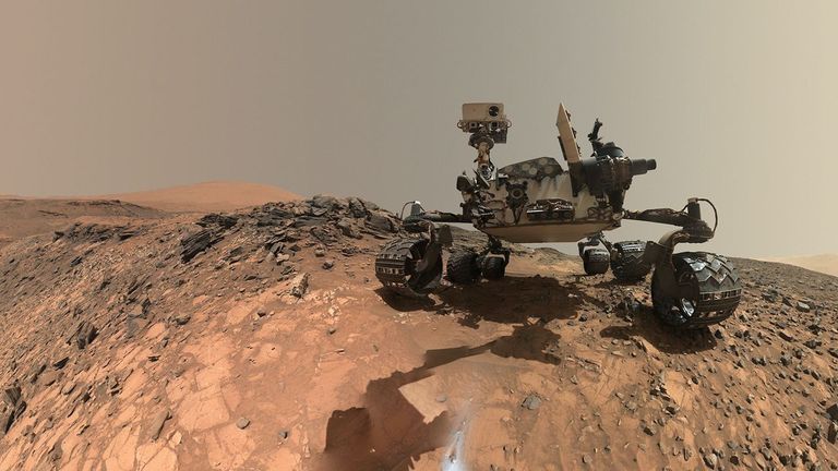 Curiosity Rover Targets 'Nazca' Site for New Mars Drilling Campaign 2 A rover sits on the hilly, orange Martian surface beneath a flat grey sky, surrounded by chunks of rock.