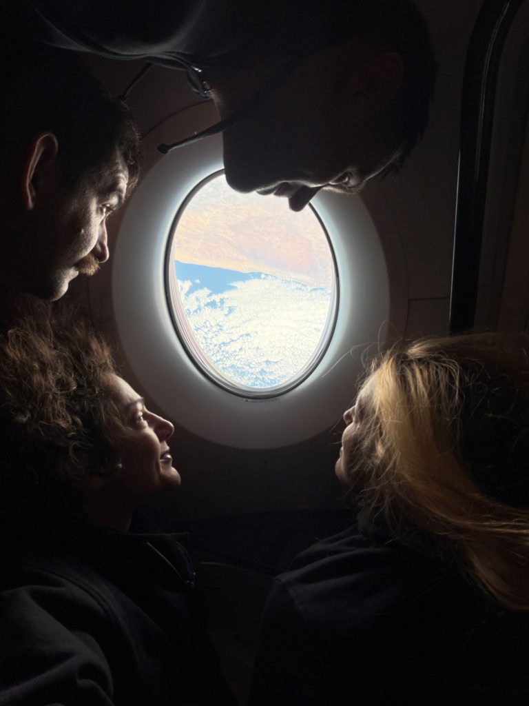 NASA’s SpaceX Crew-12 crew members surround an oval-shaped window aboard a SpaceX Dragon, looking out at clouds above blue water and a tan landmass. From the left, rotating clockwise around the image are NASA astronaut Jack Hathaway, Roscosmos cosmonaut Andrey Fedyaev, ESA (European Space Agency) astronaut Sophie Adenot, and NASA astronaut Jessica Meir. Their faces are illuminated by sunlight streaming through the window.