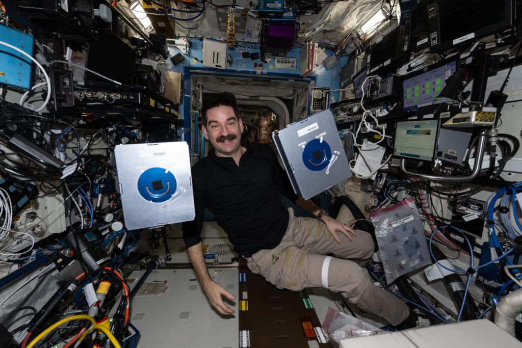 NASA astronaut Jack Hathaway floats and smiles at the camera, his face framed by two metallic silver boxes. Around him, the International Space Station’s walls are densely packed with wires, tubes, computer screens, and other hardware.